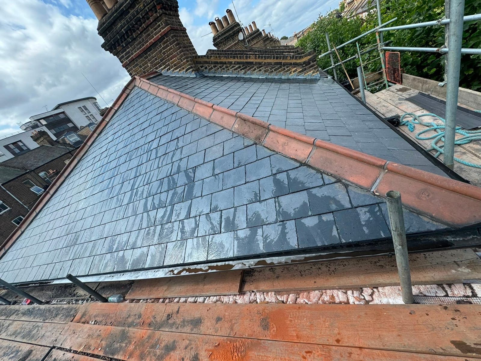 Wet freshly laid slate roof with terracotta ridge tiles and a lead-flashed chimney, scaffold alongside