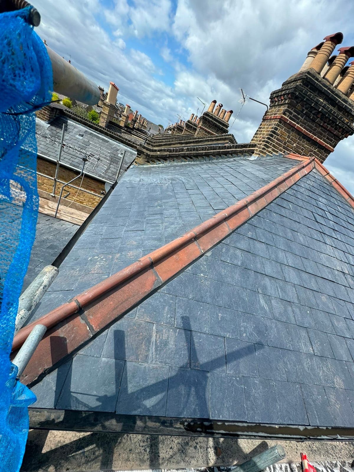Close view of a new slate roof with a bedded terracotta ridge, brick chimneys and aerials in the background